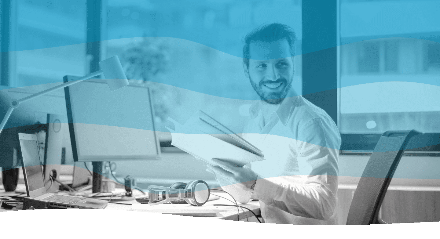 A man sits at a modern office desk with multiple computer monitors, holding open a book and smiling, framed by a large window overlooking a cityscape, with a blue overlay effect.