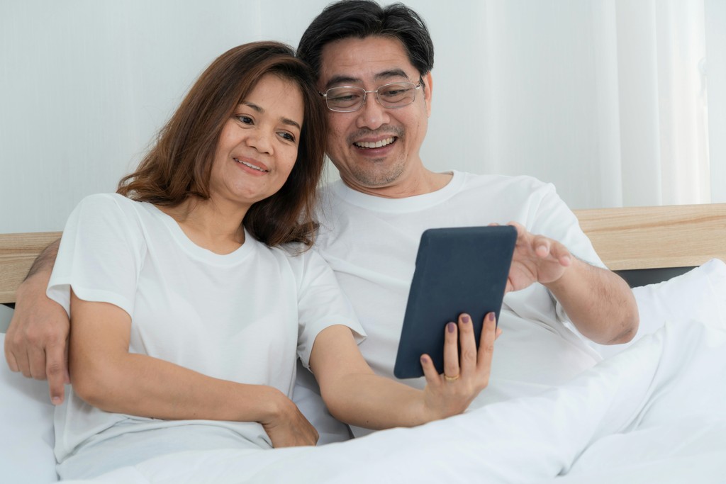 A happy middle-aged couple sits comfortably in bed, smiling as they look at a tablet together, reflecting the joy of shared digital experiences and the ease of staying connected from home.