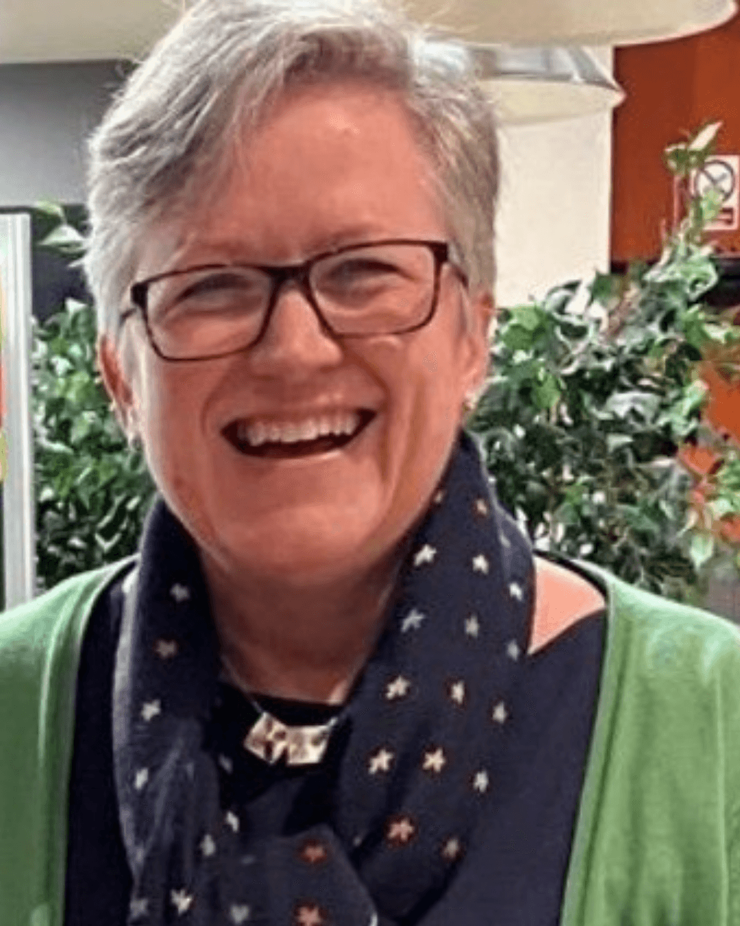 A smiling person with curly hair, wearing a leather jacket, stands in a cozy indoor setting.