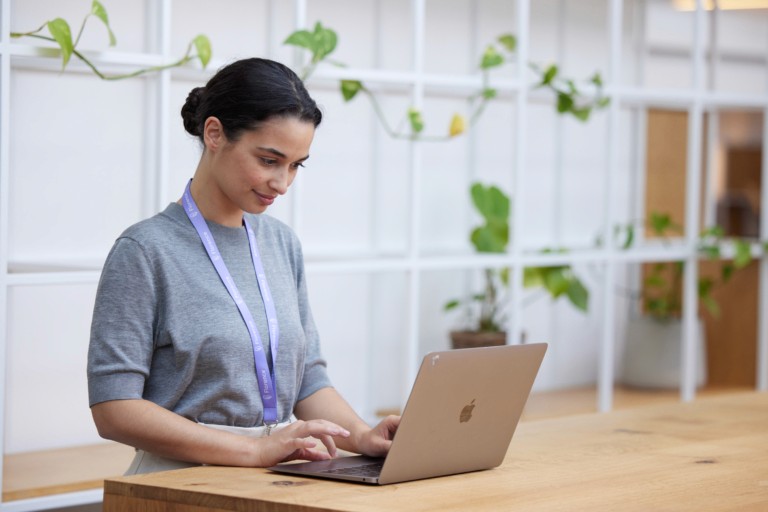A woman wearing a lanyard working on a laptop at a desk in a bright office space with plants in the background