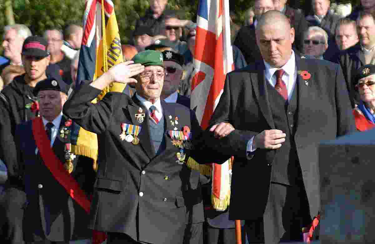 A photograph at a remembrance day event, many people in military dress in the background and in the foreground a man in a suit with short hair, wearing a poppy on his lapel stands arm in arm with an older man wearing a green beret and many medals, he is saluting
