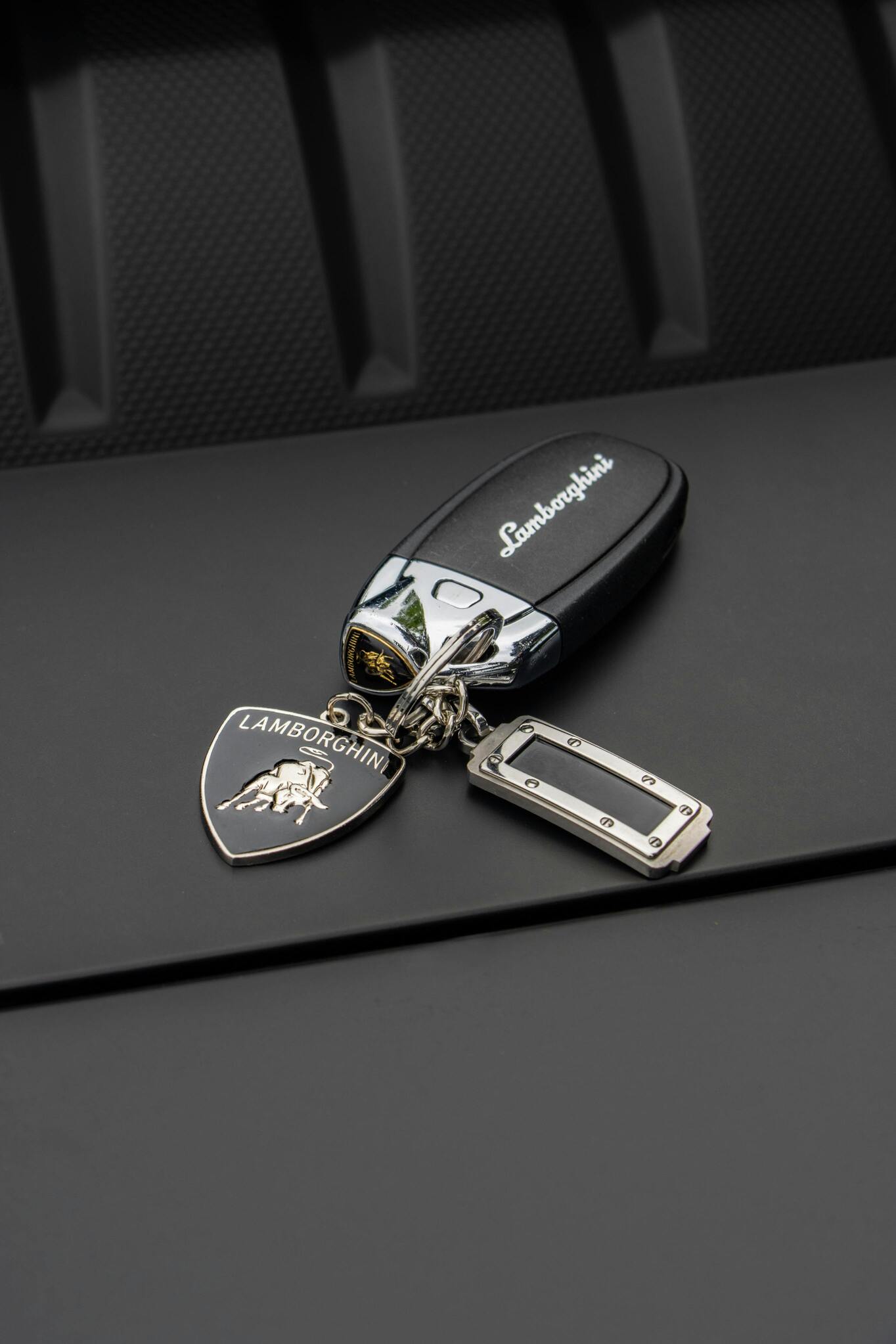 A sleek black Lamborghini car key with an attached silver Lamborghini emblem keychain rests on a textured dark surface, symbolizing luxury and automotive excellence.
