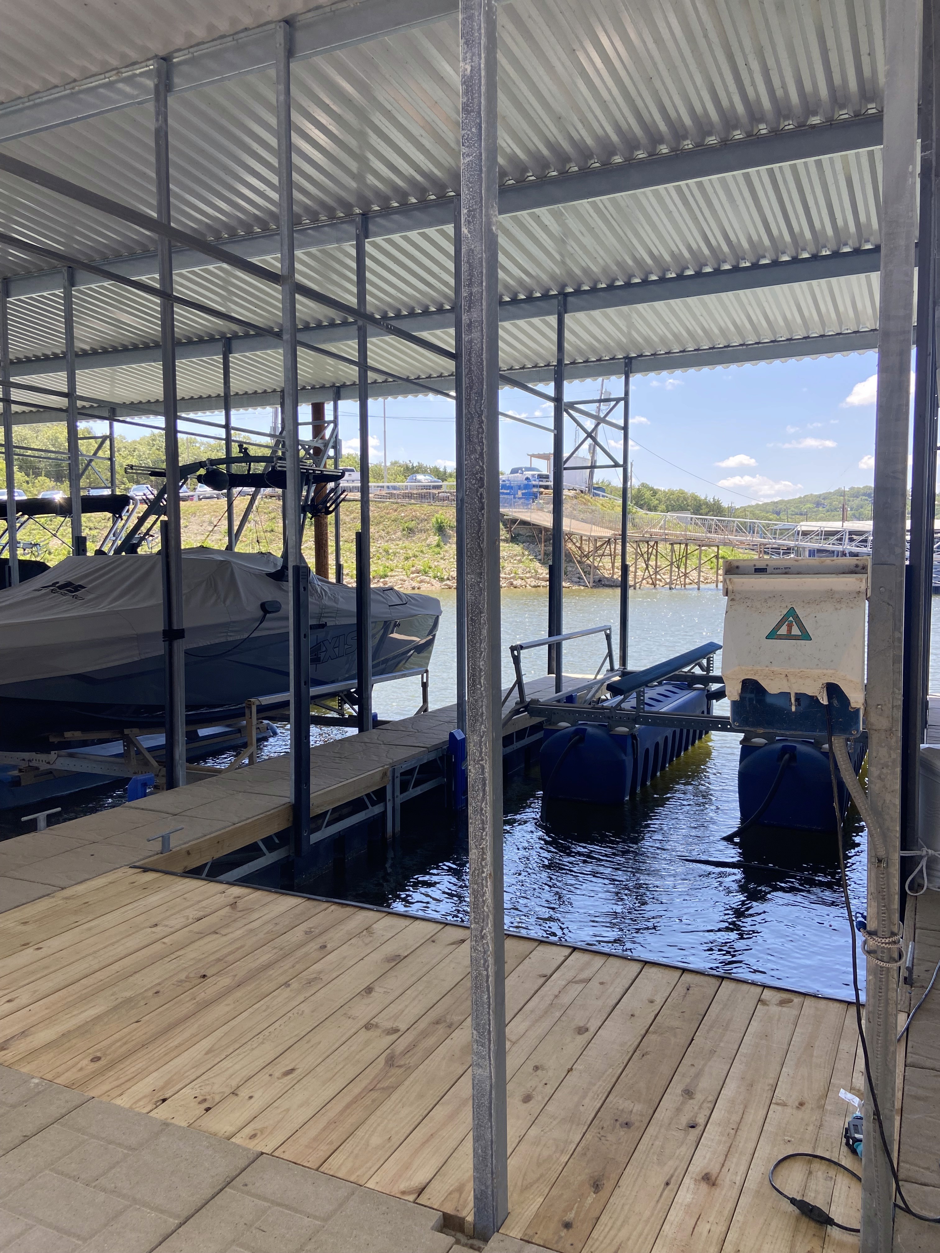 A covered boat dock with a motorboat resting in its berth, surrounded by calm water beneath a metal roof, while a wooden walkway and a distant bridge are visible against a backdrop of clear blue sky.