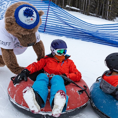 บุคคลหนึ่งกำลังลื่นไถลบนหิมะในเครื่องแต่งกายฤดูหนาว มีปฏิสัมพันธ์กับมาสคอตหมี อีกคนหนึ่งกำลังลื่นไถลอยู่ใกล้ๆ