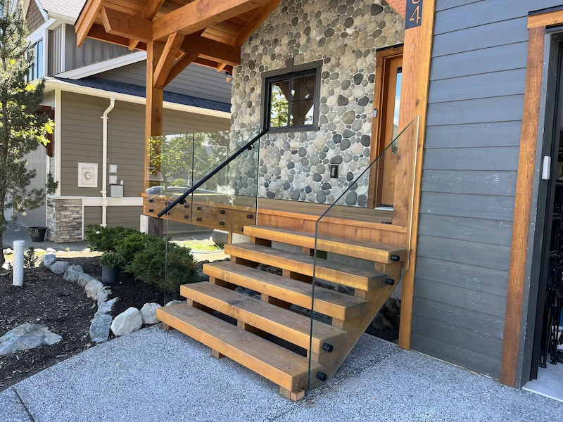 Front entry staircase with glass railing and exposed timber steps leading to a stone-accented home exterior in Burnaby, designed for strength and style.