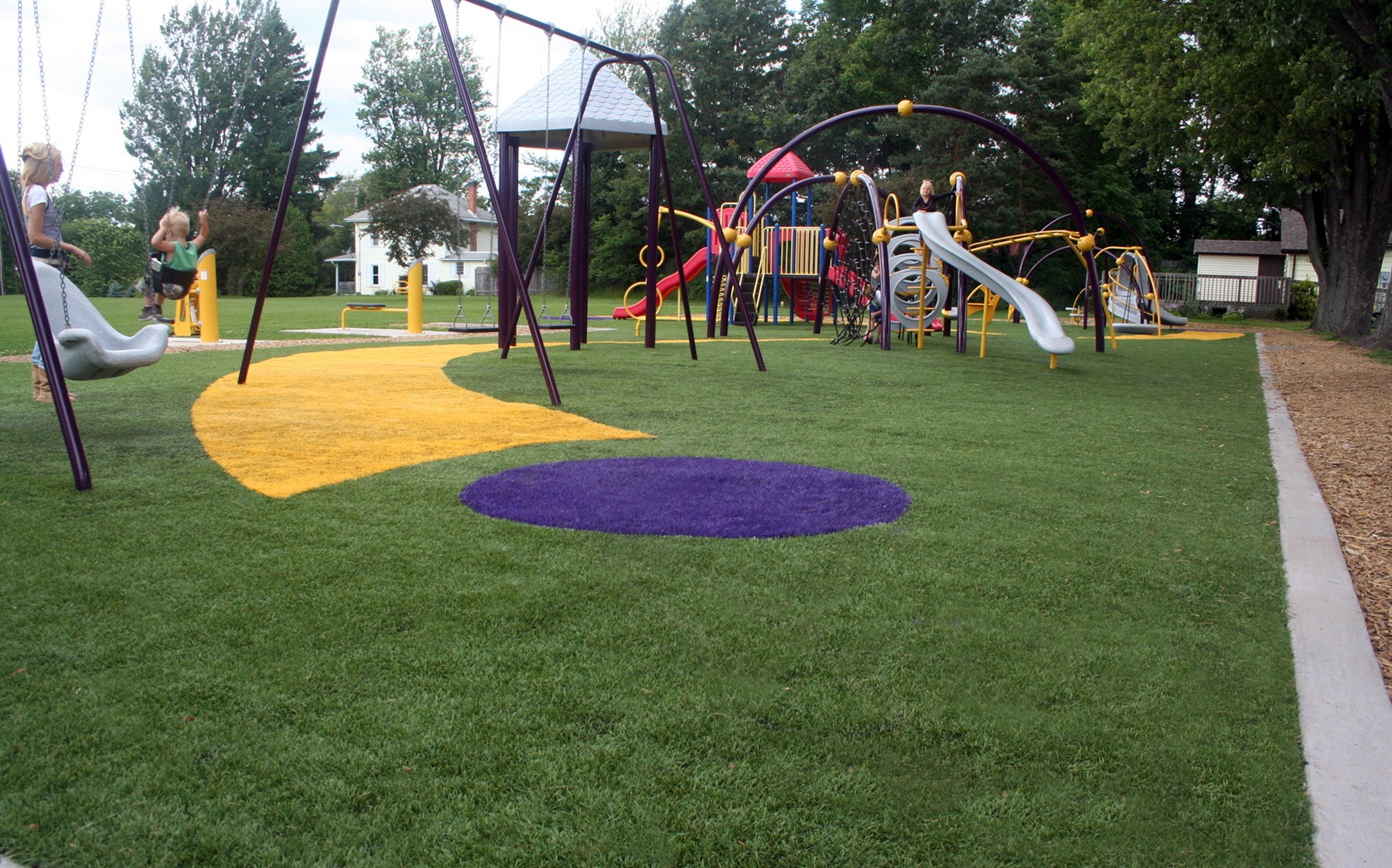 This fun-filled playground, which features a bright red slide, swings, and a jungle gym, is set in a grassy artificial turf playground by AGL Grass, perfect for kids to have fun.