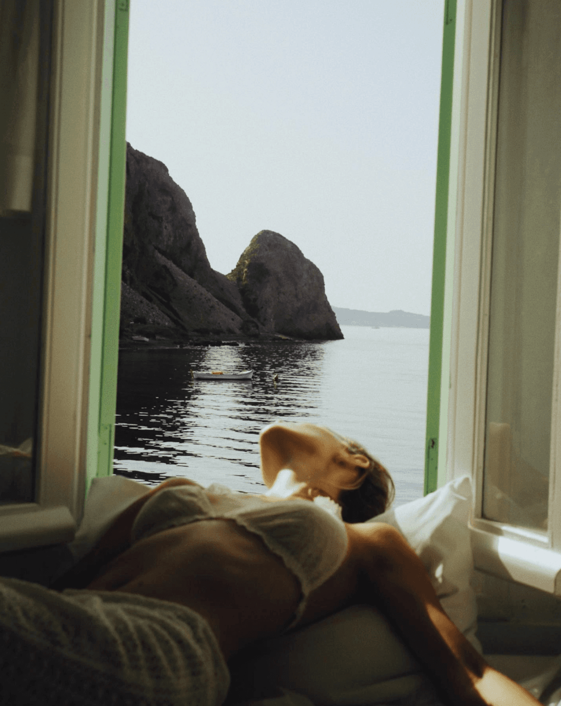 Person resting by an open window overlooking a rocky Mediterranean coastline