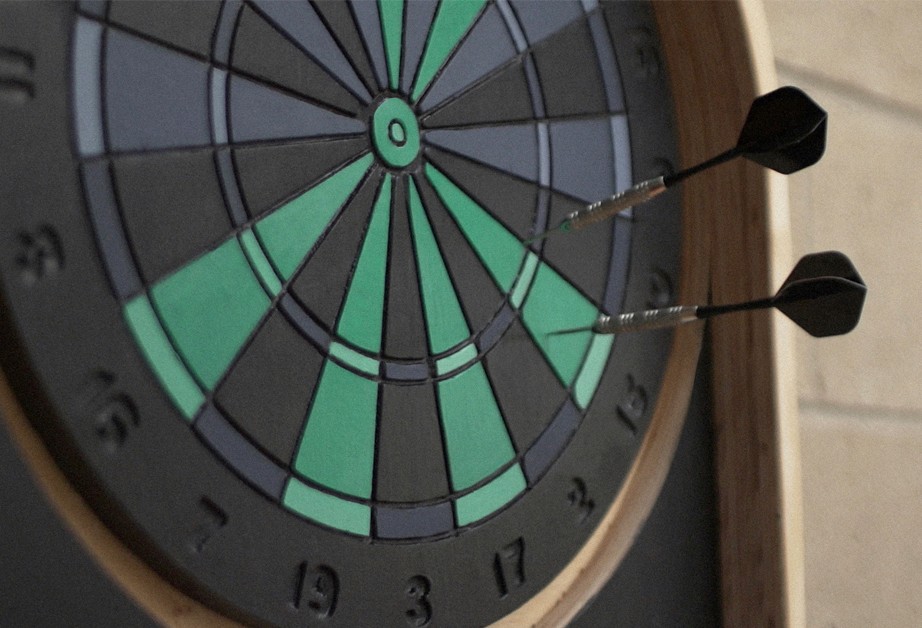 A dartboard with teal and dark gray segments is shown with two black darts embedded in the board. The board has numbers 1 through 20 around the edge. The background is a light beige wall.