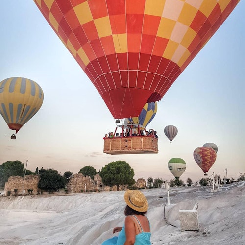 Isang babae sa asul na damit at sumbrerong gawa sa dayami ang nakamasid habang umaangat ang mga makukulay na hot air balloon sa langit sa ibabaw ng mabatong tanawin.