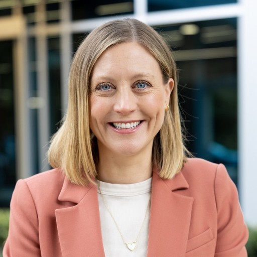 A focused woman with long red hair is seated at a desk, wearing a black blazer and dark blouse. She intently looks at camera, smiling slightly, with an open laptop in front of her. The setting is a warm and serene office setup in the background. Soft daylight filters through, creating a calm atmosphere. The composition captures her in a frontal profile with a clear emphasis on her concentration and the serene indoor setting.