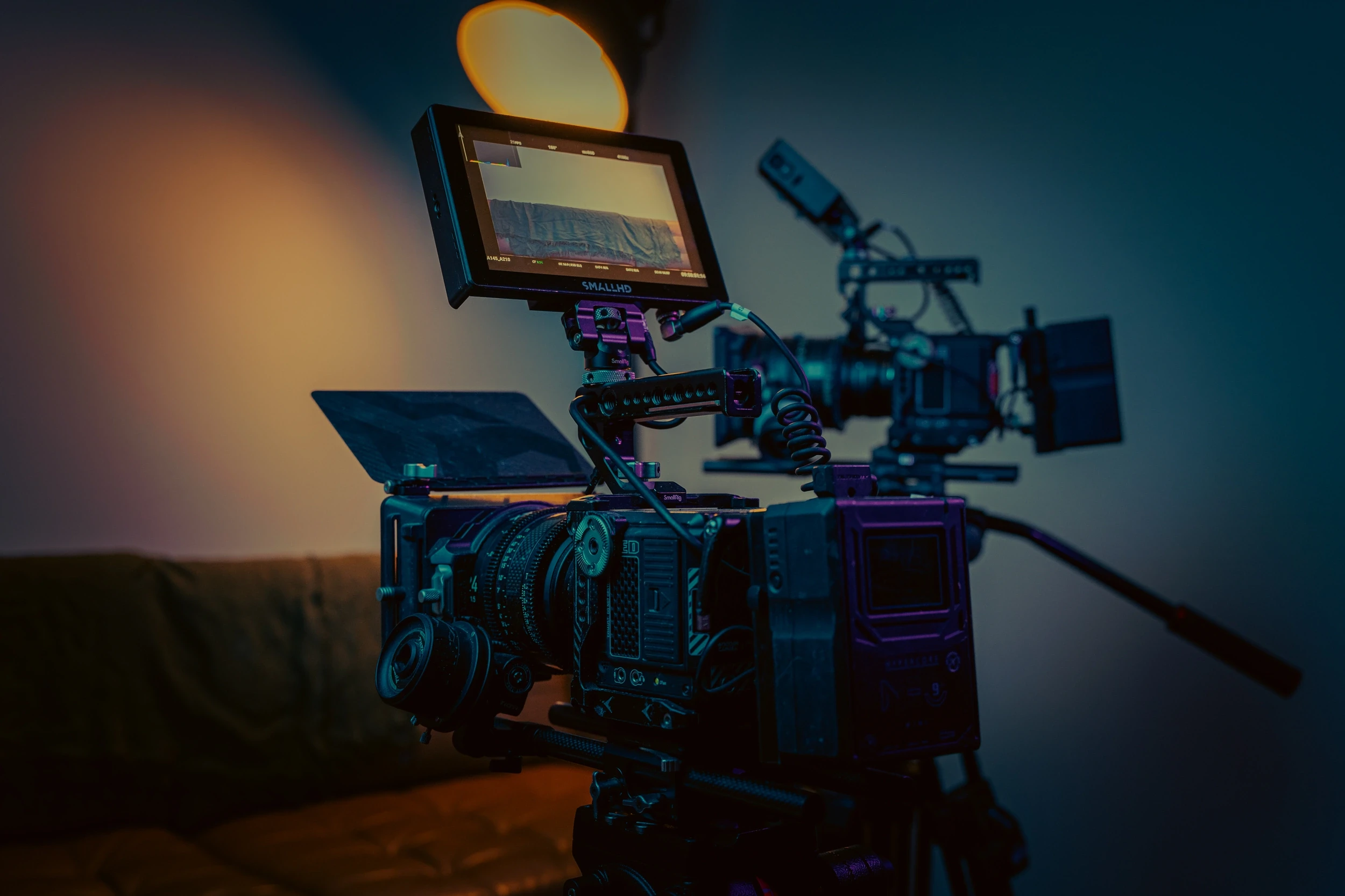 Cinema camera built up on a tripod with an on-board monitor and boom microphone, lit warm against blue