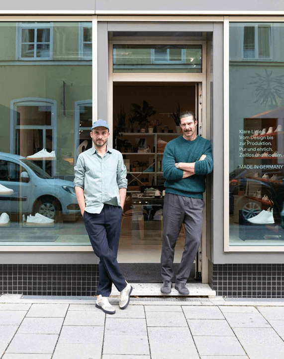 Two people stand casually in the doorway of a modern shop with large glass windows, displaying interior decor elements and a reflection of a parked car on the street.