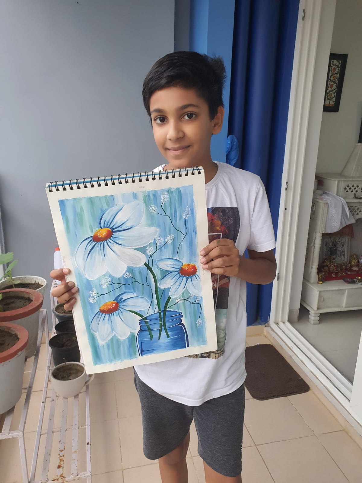 A boy holds a colorful artwork featuring flowers in shades of blue and white, smiling, standing in the Creative Pod studio.