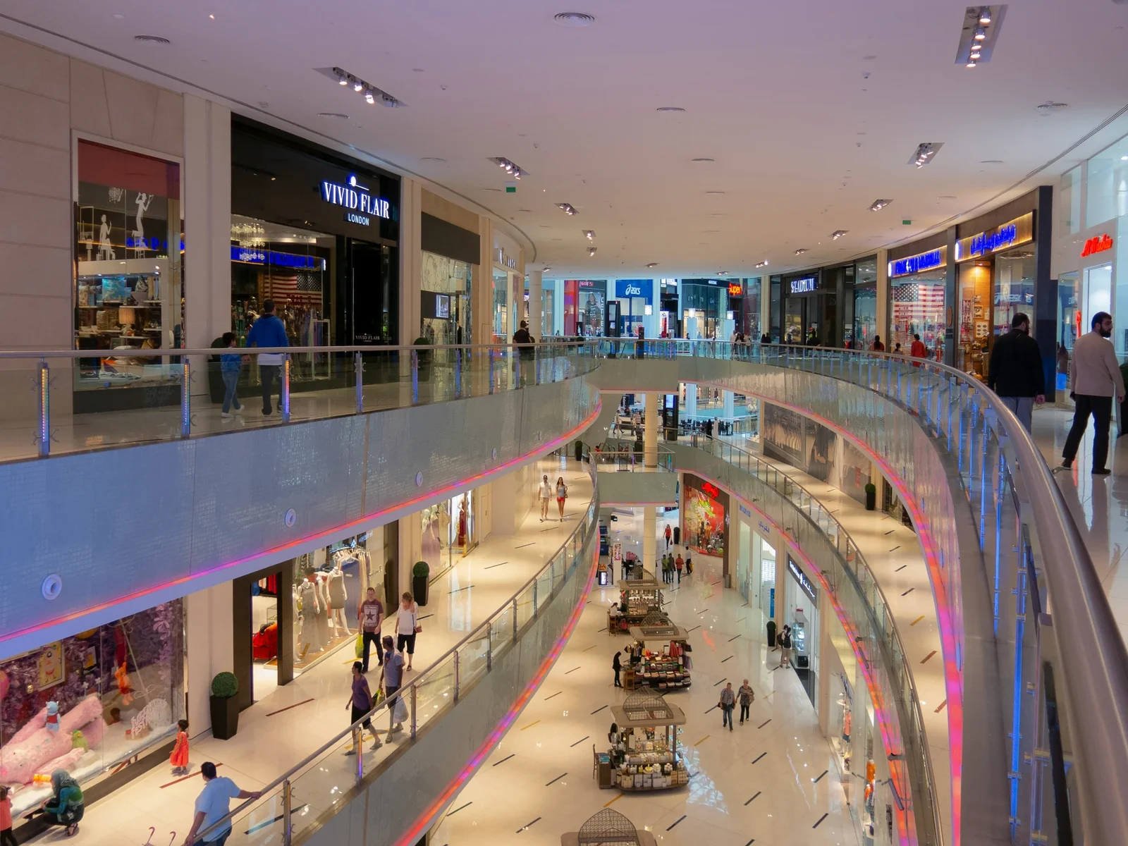 Multi-level shopping mall interior with stores, shoppers, and neon-lit railings displaying retail storefronts including Vivid Flair London.