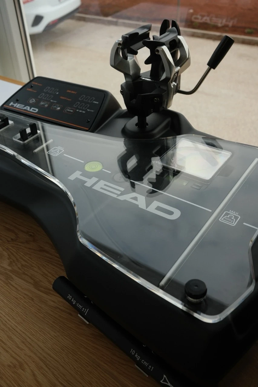 Professionelle Besaitungsmaschine der Marke HEAD, platziert auf einer Holzoberfläche. Die Maschine verfügt über eine digitale Anzeige, eine robuste Schlägerhalterung mit metallischen Klemmen und eine transparente Abdeckung mit dem HEAD-Logo. Im Hintergrund ist ein Fenster mit Blick auf einen Parkplatz sichtbar.