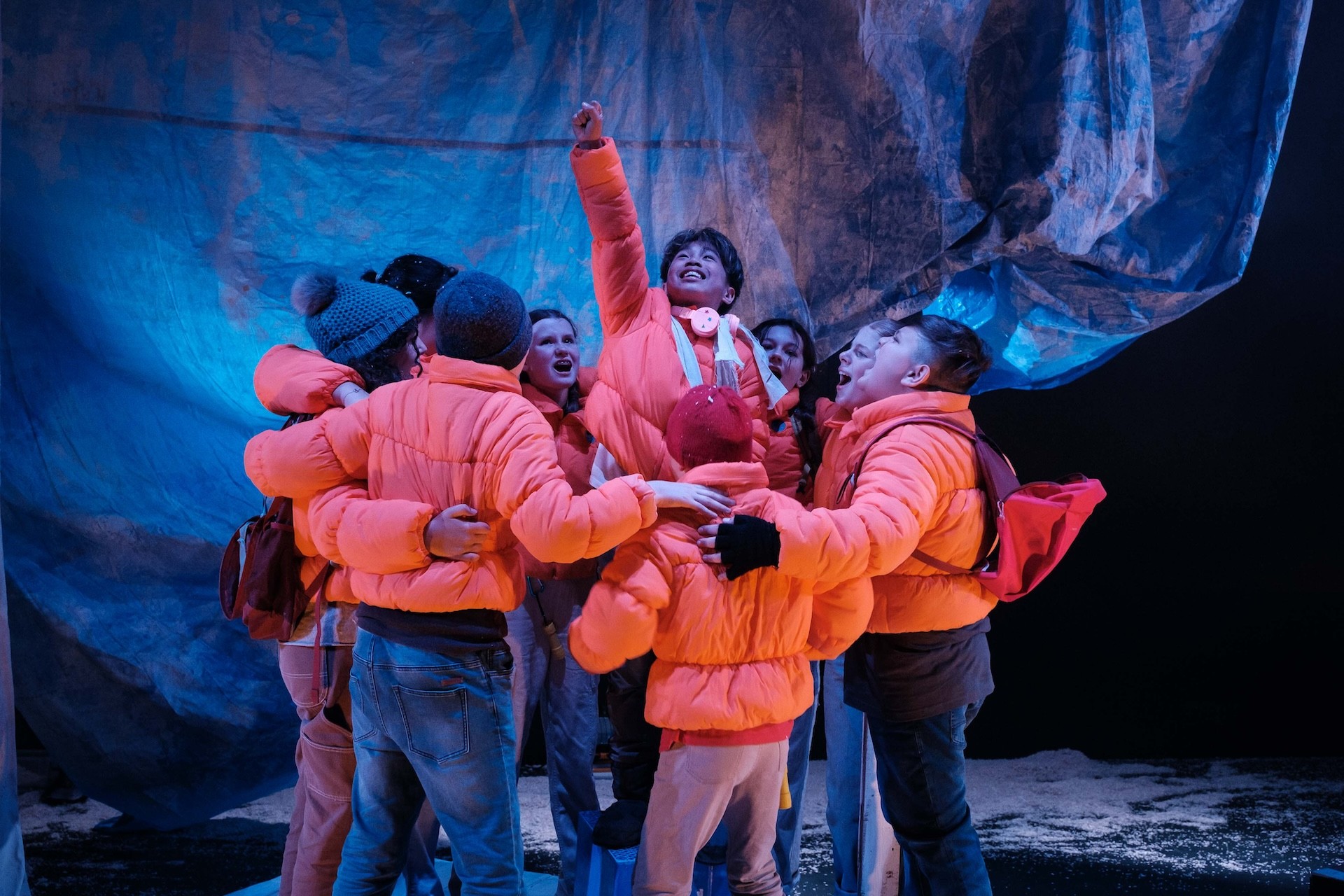 A group of performers huddle together in their orange jackets, cheering as one performer in the centre is lifted slightly, pointing triumphantly upward under blue stage lighting.