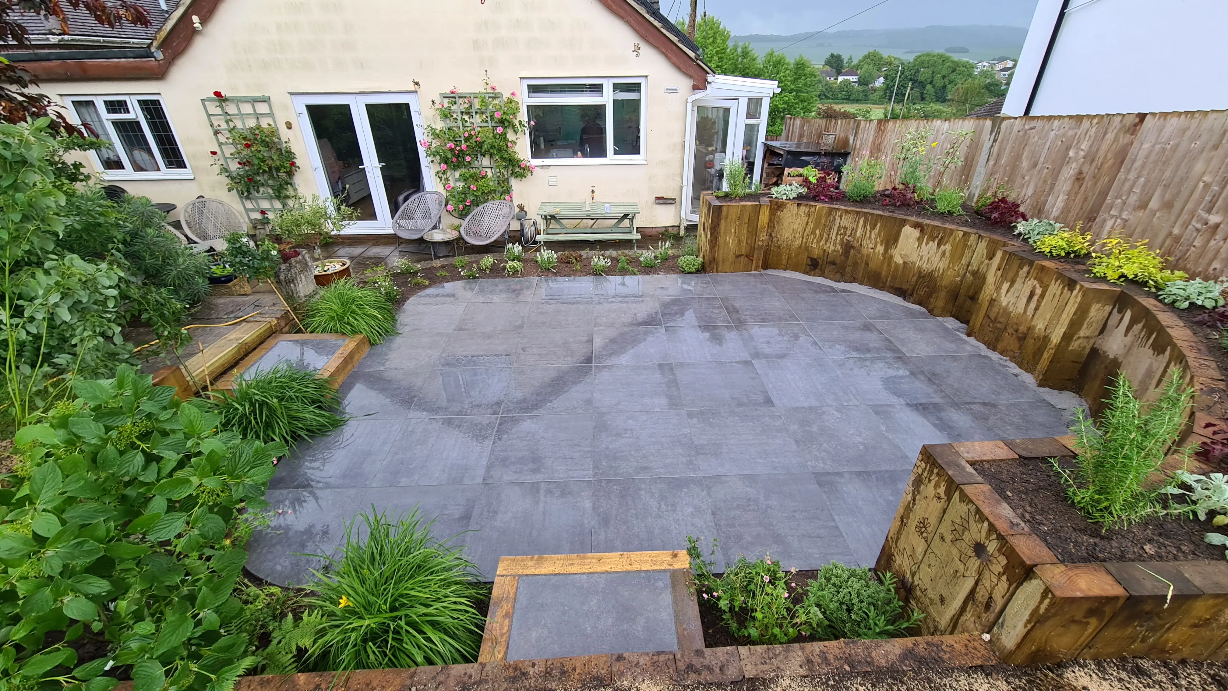 A landscaped backyard with a circular stone patio, bordered by wooden fencing and lush greenery.