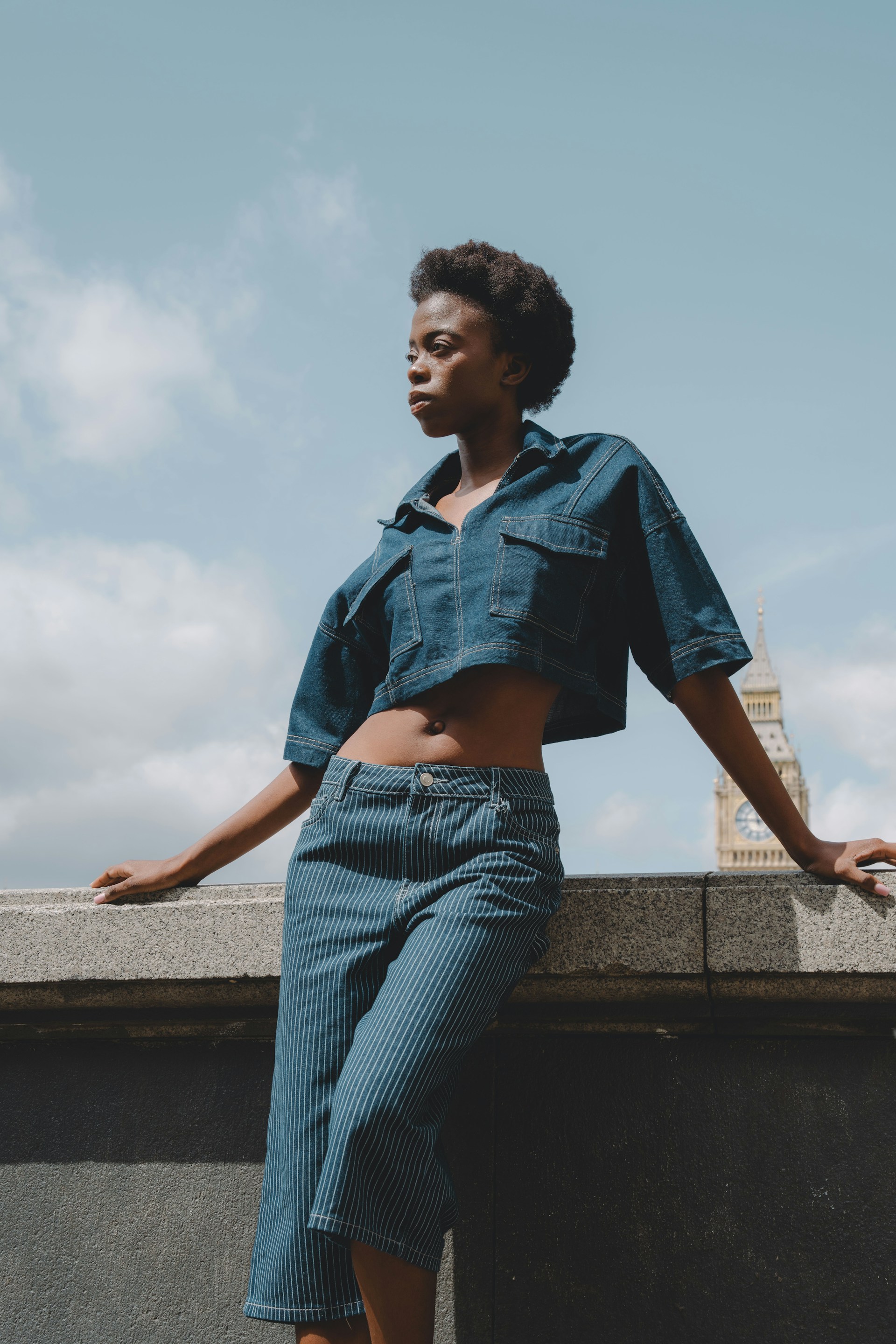 A portrait of a woman with short natural hair posing outdoors. She is wearing a cropped denim shirt and pinstriped denim trousers, leaning against a stone wall with the Big Ben clock tower visible in the background.