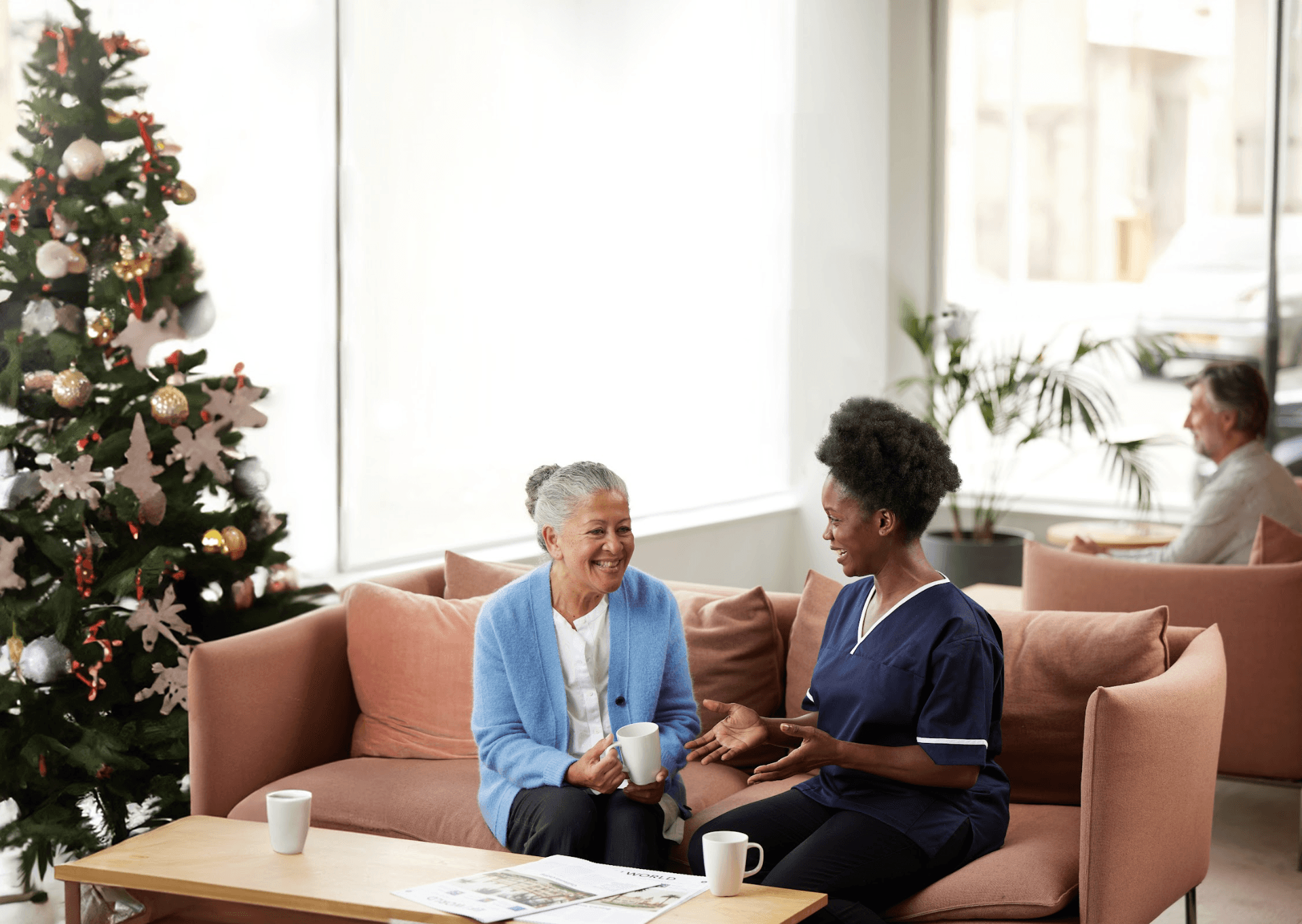 An elderly woman and a care worker in nursing uniform sitting together on a sofa, smiling and chatting near a decorated Christmas tree