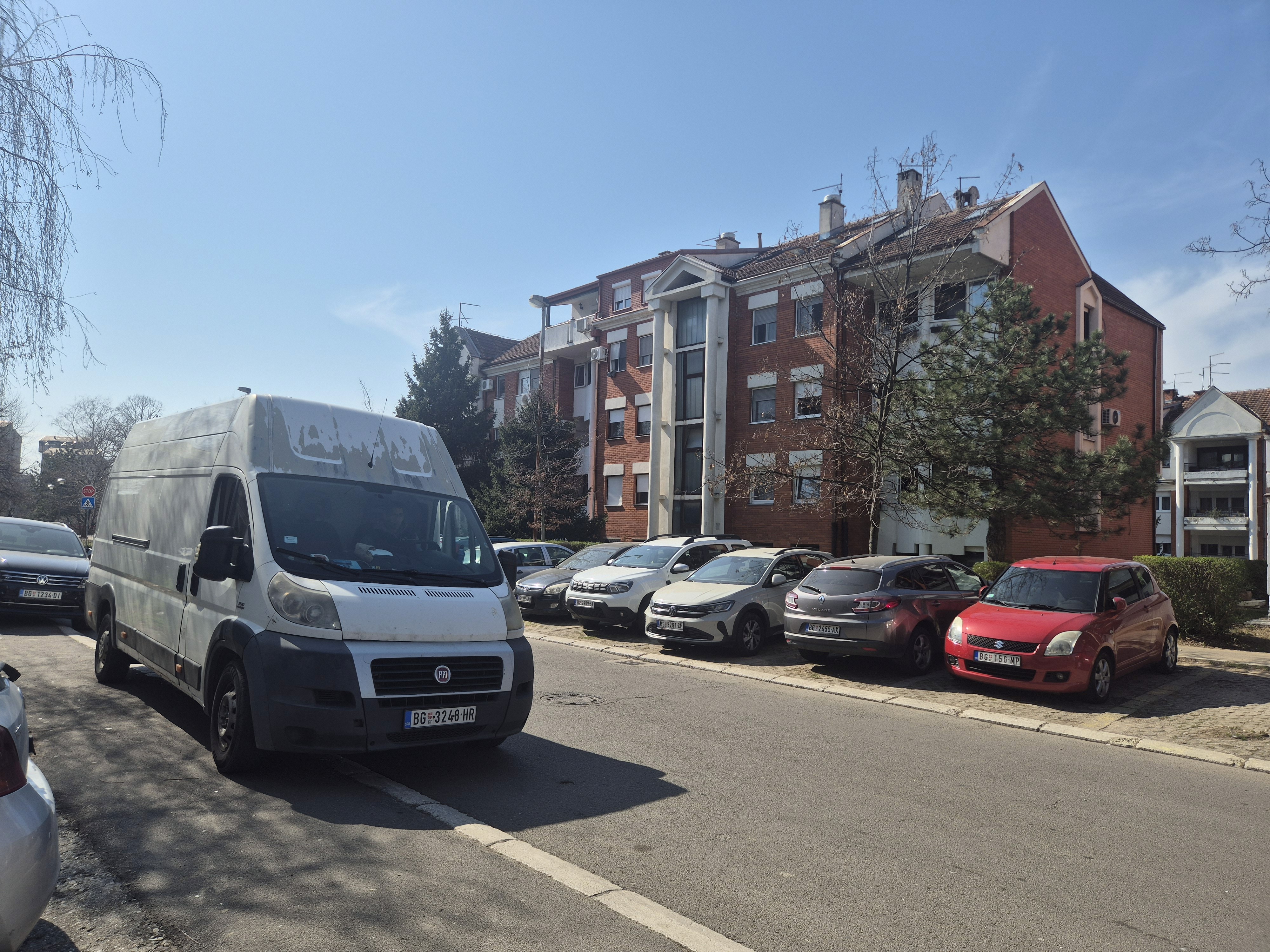 Beli kombi parkiran uz trotoar u mirnoj gradskoj ulici, ispred stambene zgrade od crvene cigle, pod vedrim nebom.