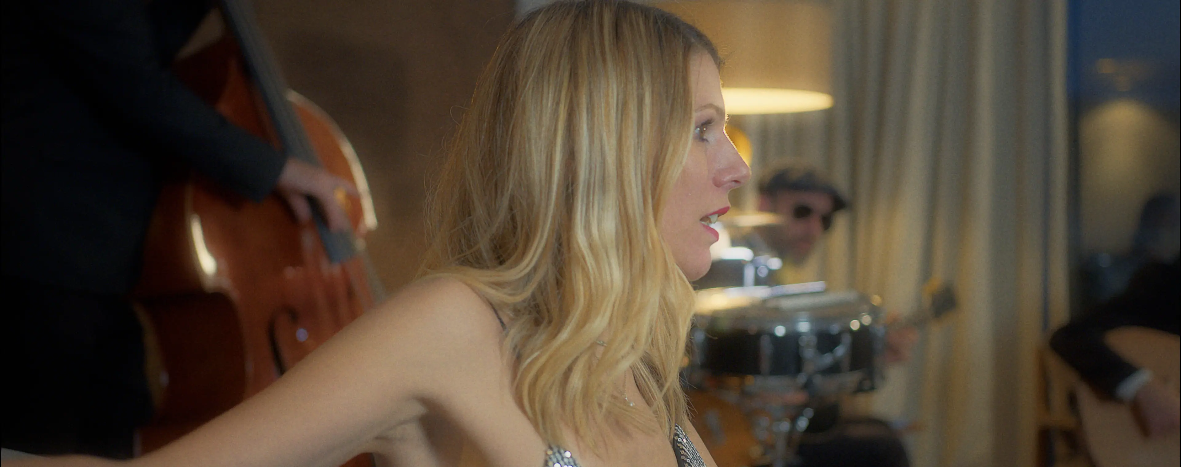 A woman with long blonde hair is singing passionately while musicians in the background play a double bass, drums, and guitar in a warmly lit room.