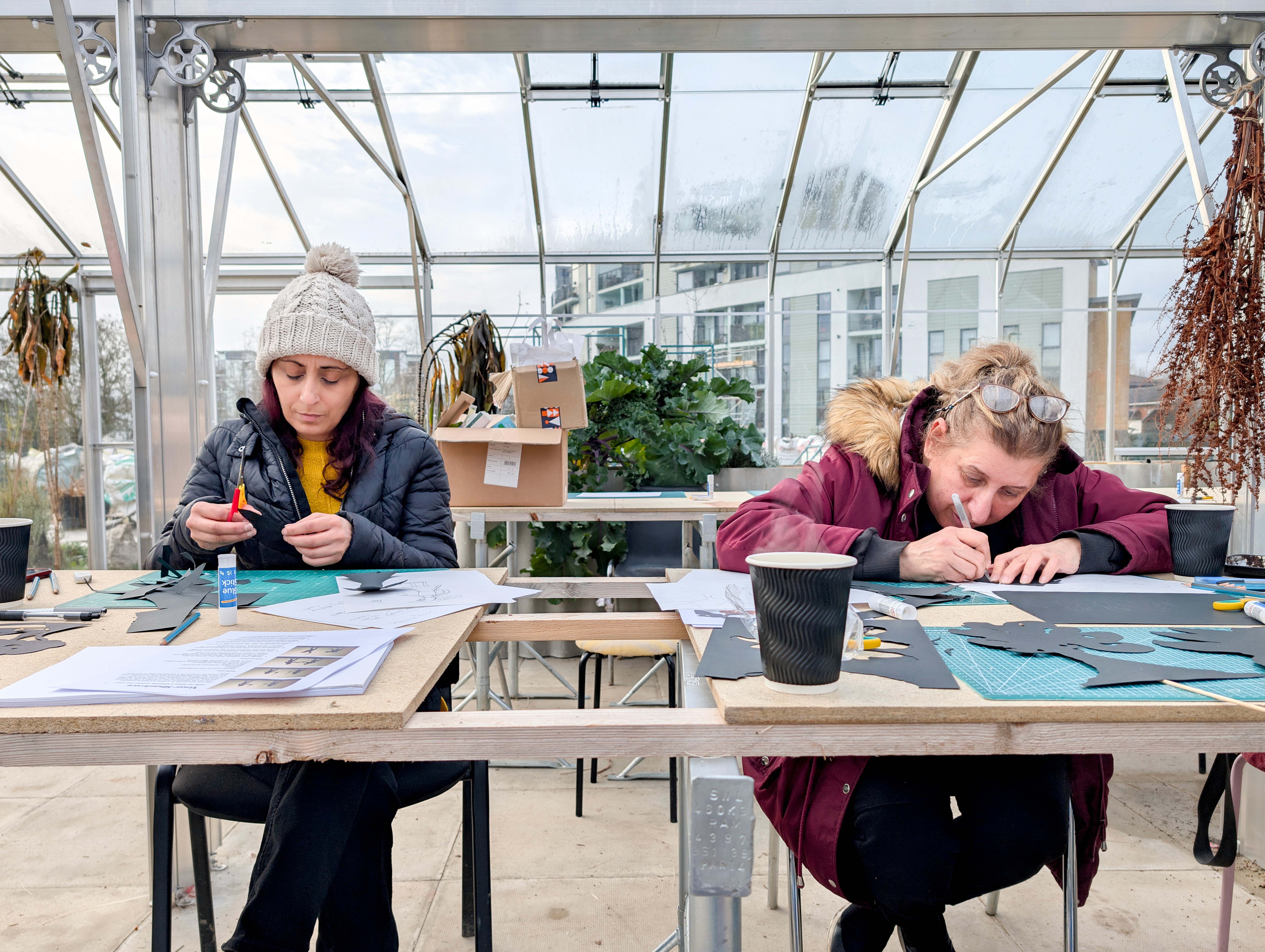 Two people seated at tables inside a glass greenhouse, focused on crafting paper cut-outs. The tables are covered with cutting mats, paper, glue, and tools, with plants and workshop materials visible in the background.