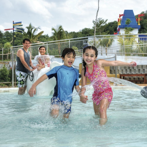 Two children play and splash in a shallow pool, while two adults look on smiling. Tropical background with slides and attractions.