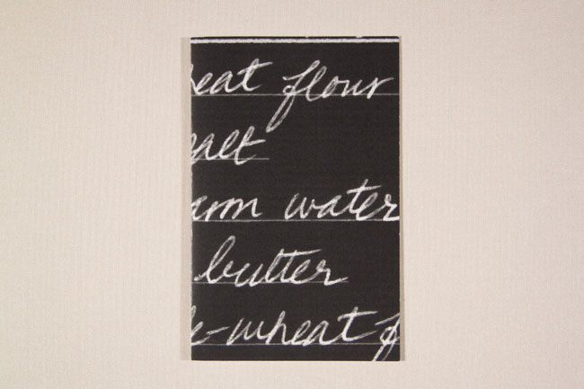 A black rectangular panel on a white wall displays white handwritten text. The text reads, from top to bottom: “heat flour”, “heat”, “arm water”, “butter”, and “wheat”. The text is centered and written in a cursive style.