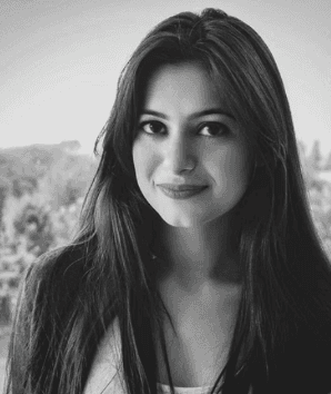 A smiling woman with her arms crossed, standing against a dark green background. She has long, dark hair.