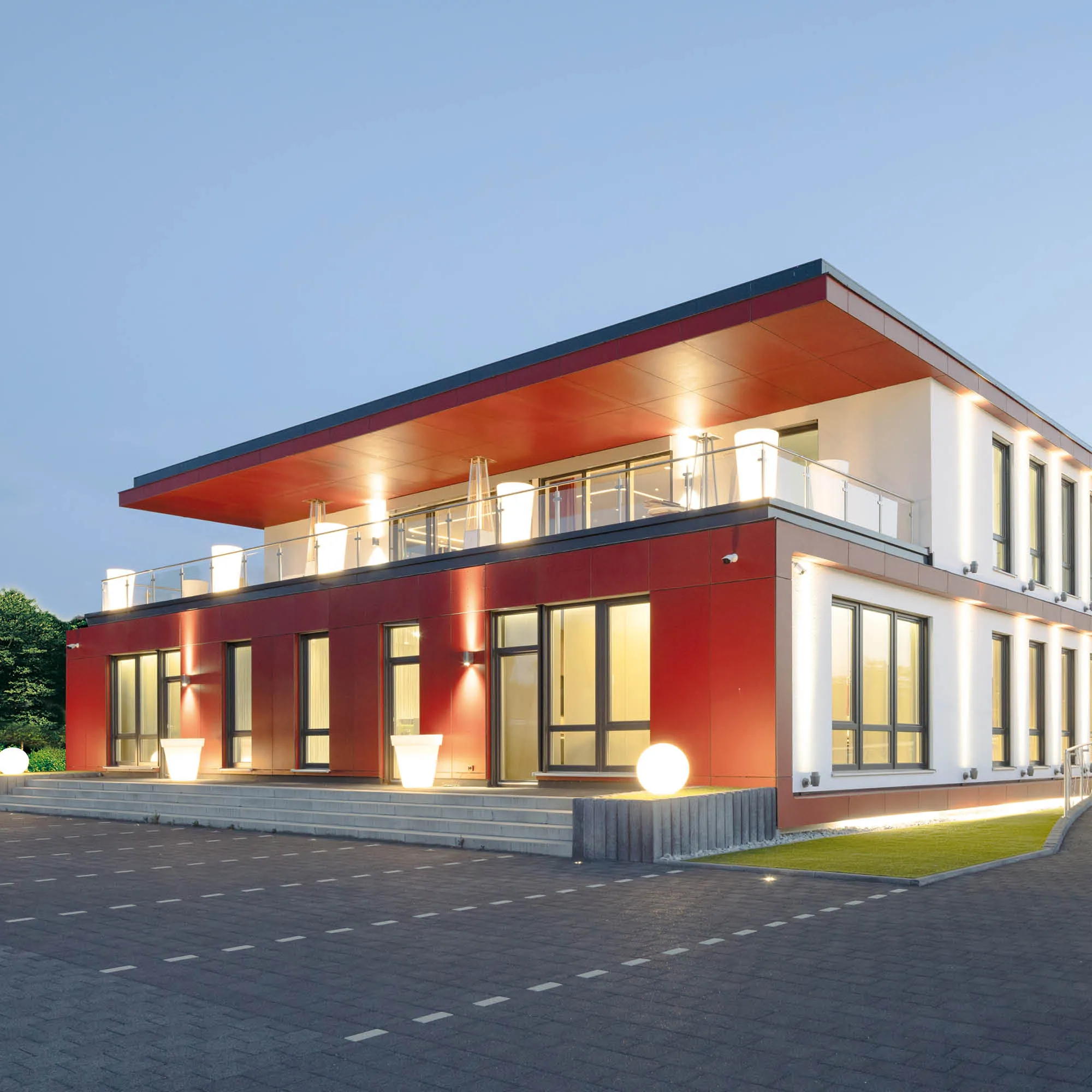 Modernes Bürogebäude mit roten Akzenten, Glasbalkonen und Außenbeleuchtung bei Dämmerung.