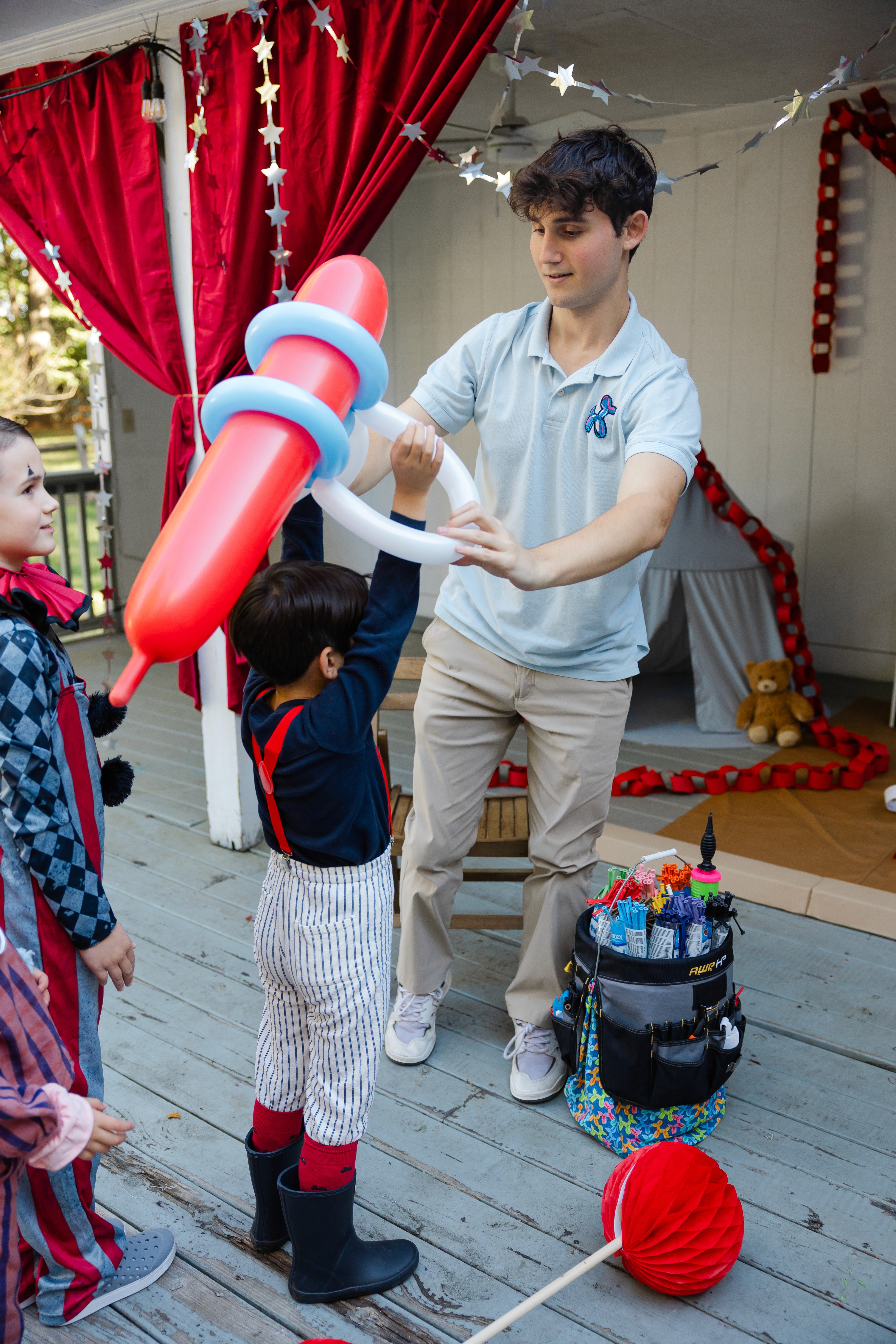 A balloon artist helps a young child put on a red and white balloon jetpack while other children look on in excitement. The scene is set on a wooden deck, with colorful decorations adding to the fun environment.