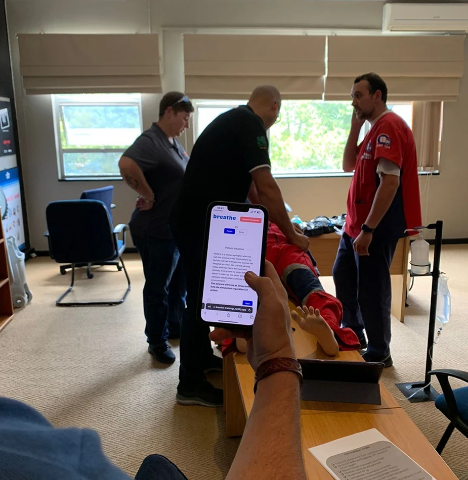 Medical training session with professionals gathered around simulation mannequin while smartphone displays Breathe app interface for emergency response.