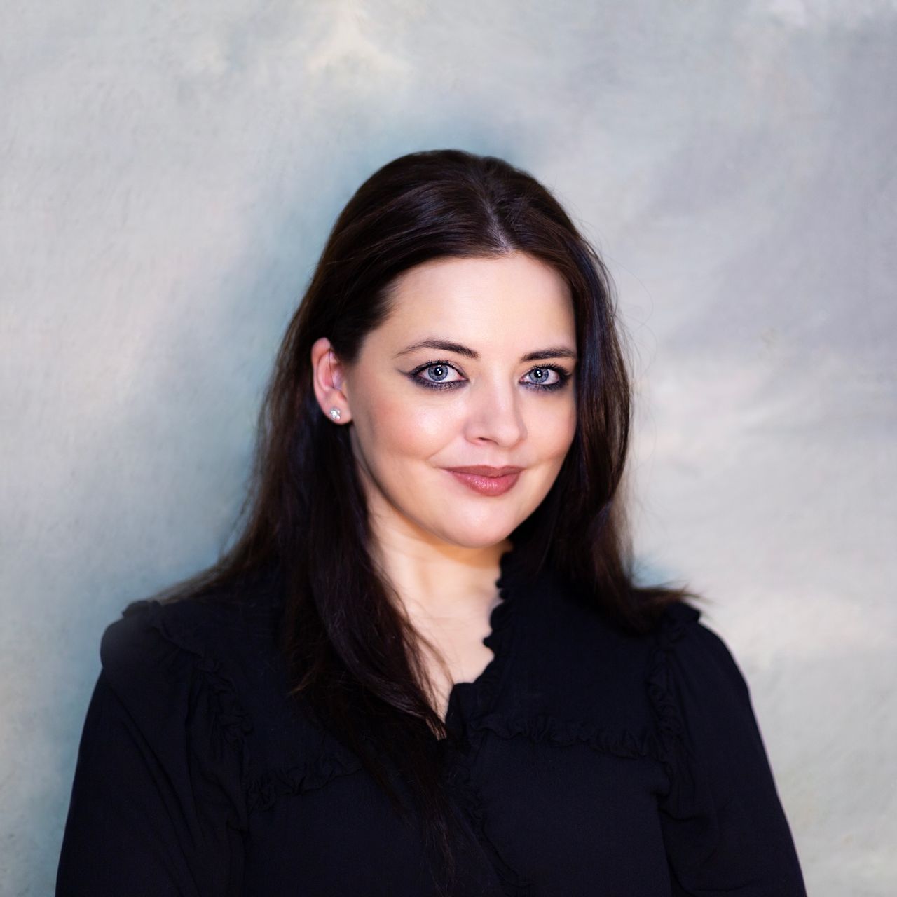 Smiling female therapist with short gray hair, wearing a brown top and a purple shawl, surrounded by a cozy background.