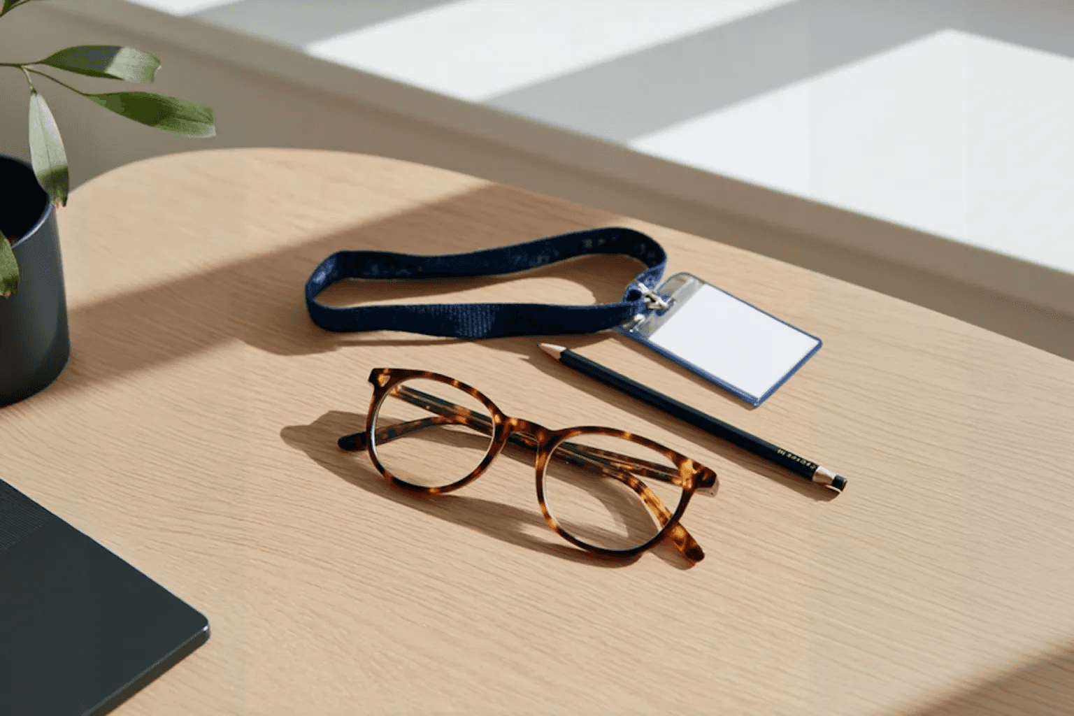 Close-up of a desk with a pair of tortoiseshell glasses, a lanyard with an ID badge, a pencil, and a notebook in soft natural light.