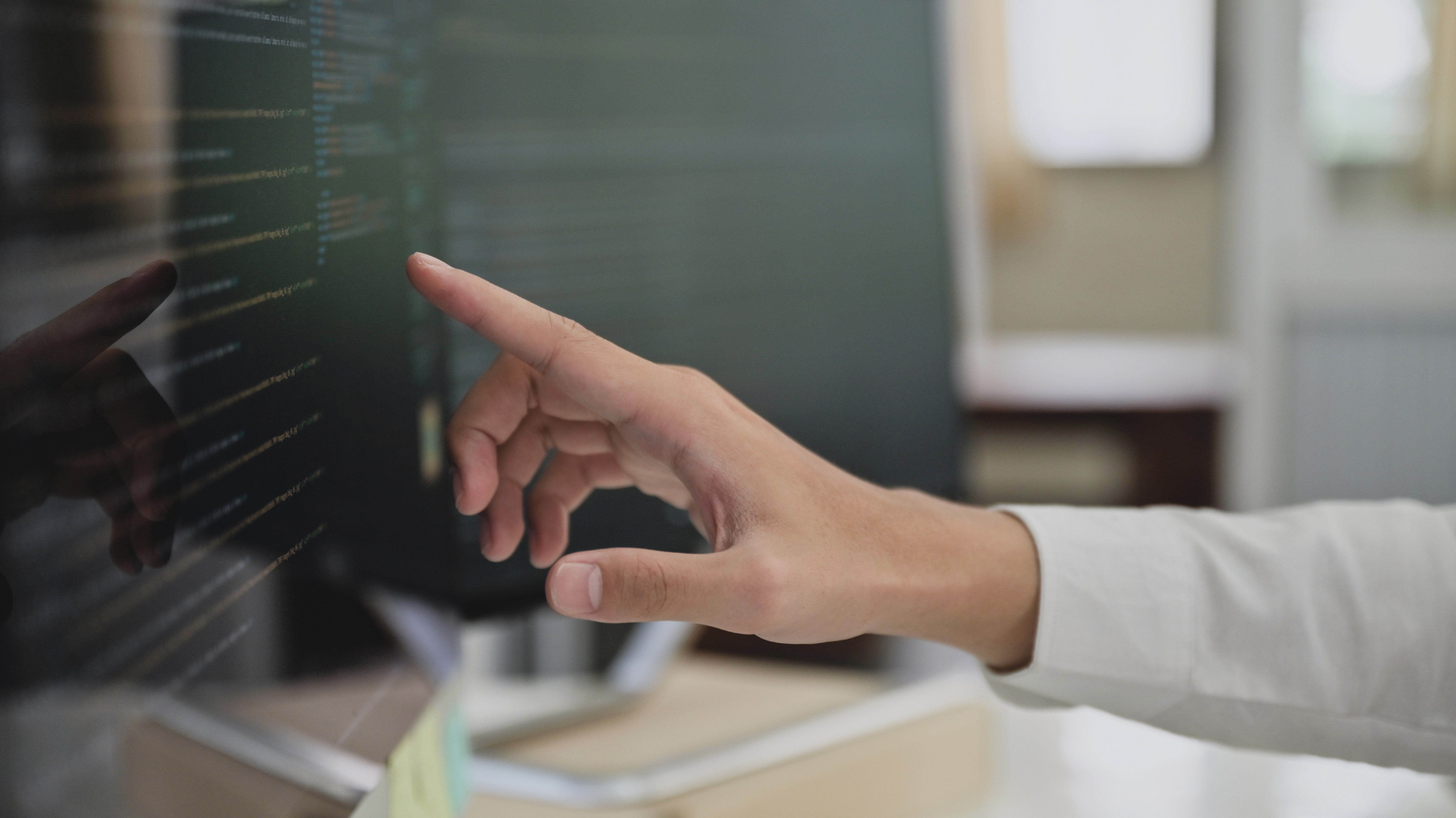 Close-up of a person pointing at code displayed on a computer screen in a modern office environment.