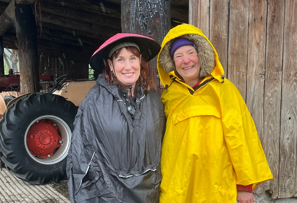 Two women smiling in rain ponchos and hats beside a barn during a rainy Fall Farm Fest.