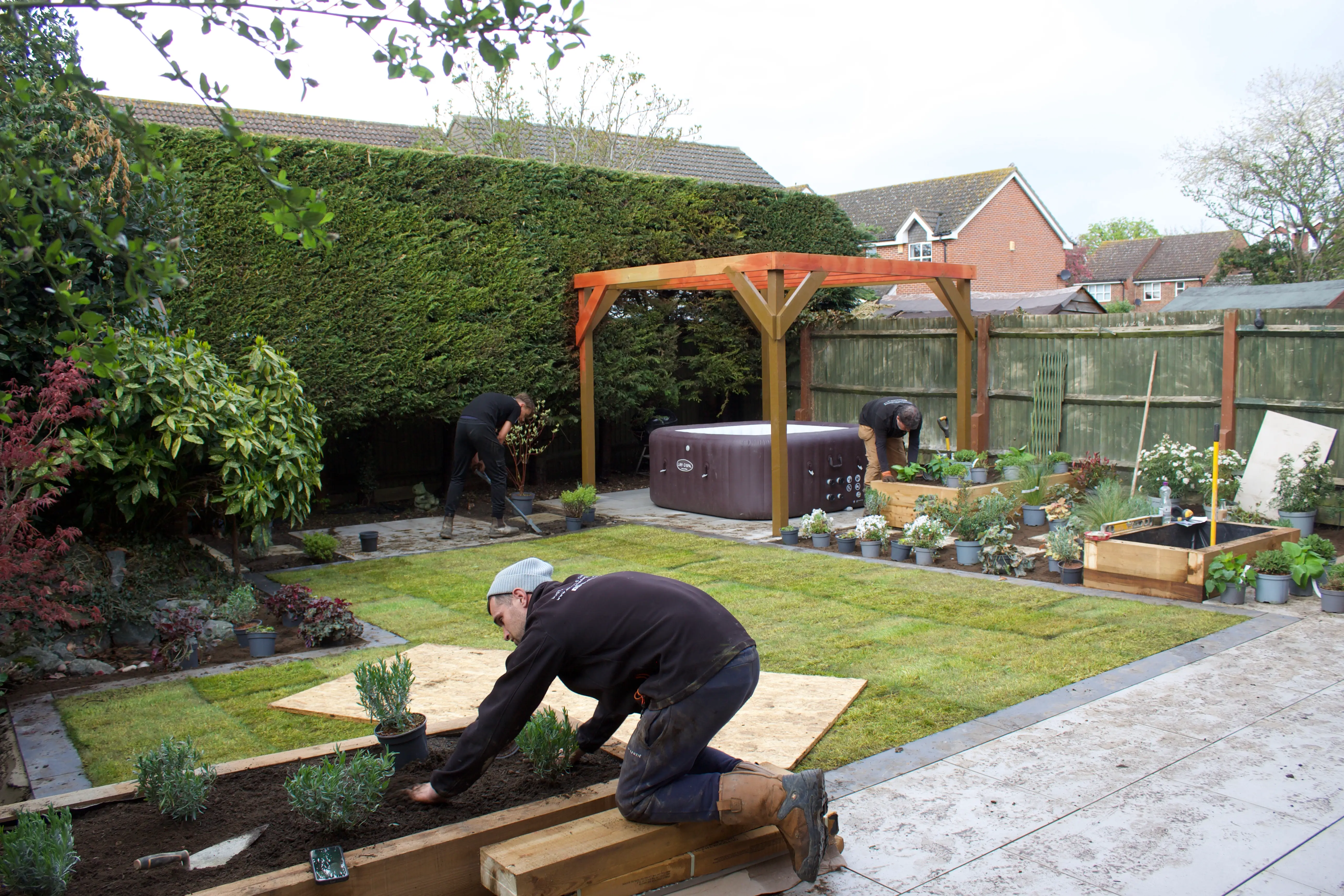 A person kneels in a garden, working on a patch of soil with plants and a gazebo in the background.