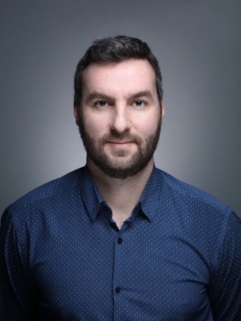 Young man with wavy hair wears a dark overcoat, rust-colored sweater, white shirt, and patterned tie, exuding a poised and sophisticated demeanor.