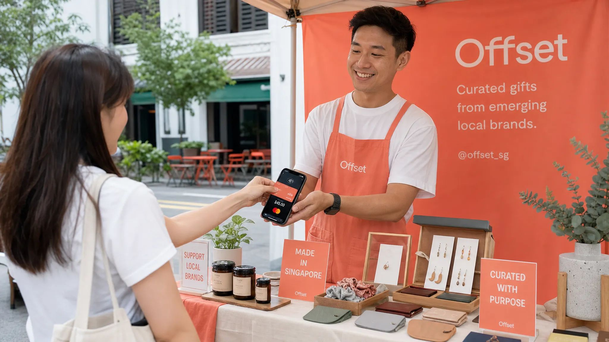 A Singapore pop-up stall with a merchant accepting a contactless card payment on a smartphone while a customer taps a card, with neatly arranged products on the table beside them.