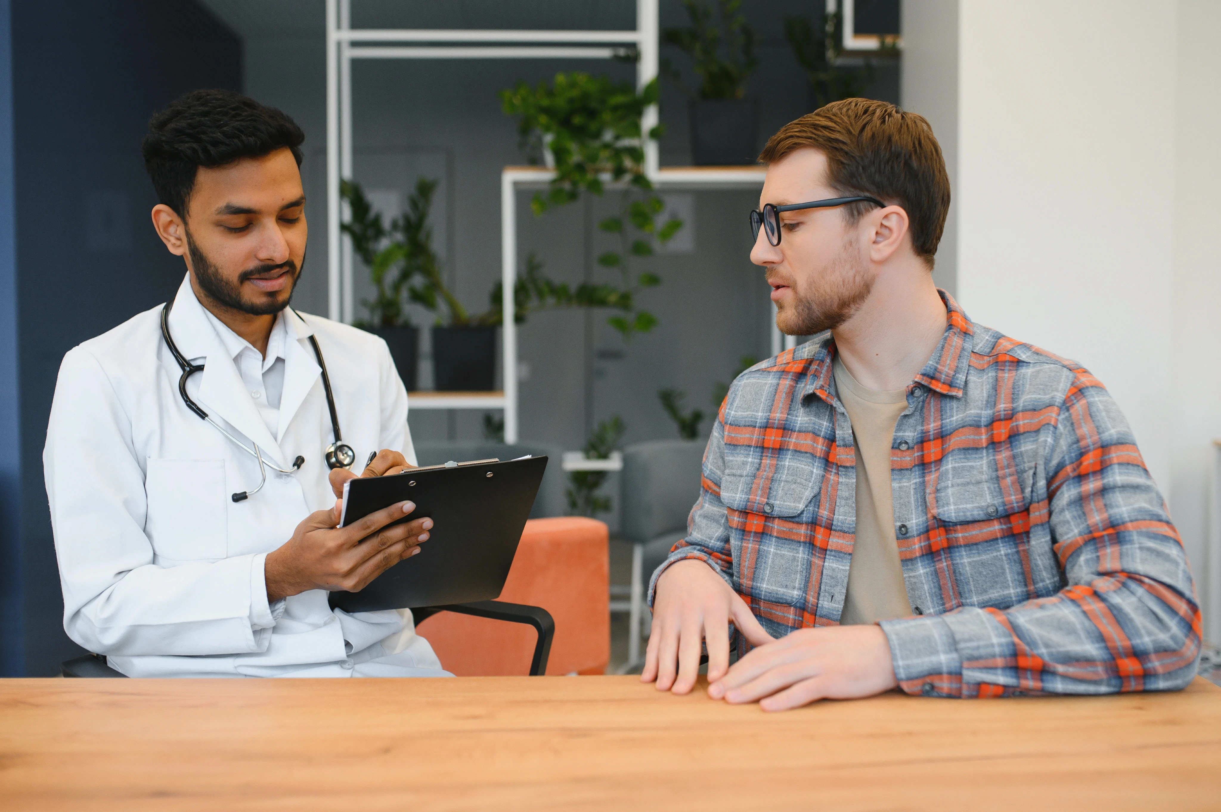 A doctor with a stethoscope holds a clipboard, attentively listening to a man in glasses and a plaid shirt. Background features green plants, conveying a calm, professional setting.