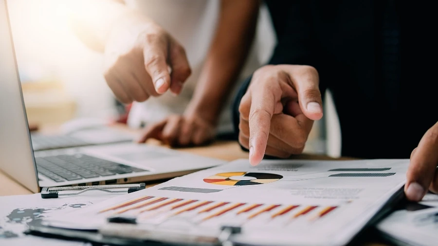 People pointing at charts and graphs on paper beside a laptop during a discussion.