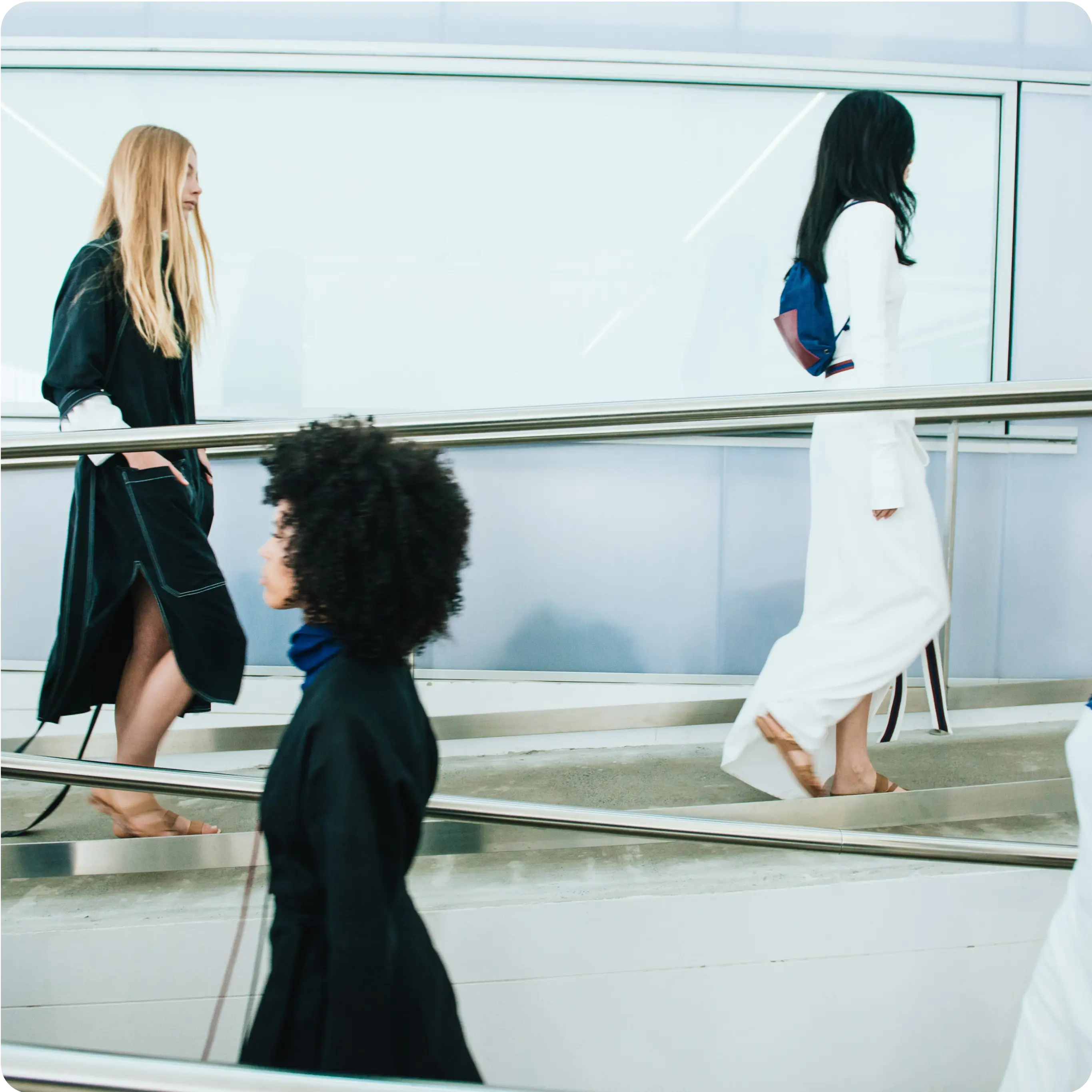 Three individuals are walking on a sloped pathway in an indoor setting. The person closest to the camera has curly black hair and is wearing a dark outfit. The middle person has long blonde hair, is barefoot, and is also dressed in dark clothing. The third person, further in the background, has long black hair, is carrying a backpack, and is dressed in white.