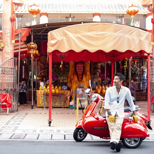 A man in light-colored clothes sits on a red scooter in front of a vibrant temple with red decorations and a large golden statue.