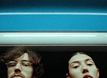 Two people appear above a blue background, partially obscured, each looking down at the camera with thoughtful expressions.