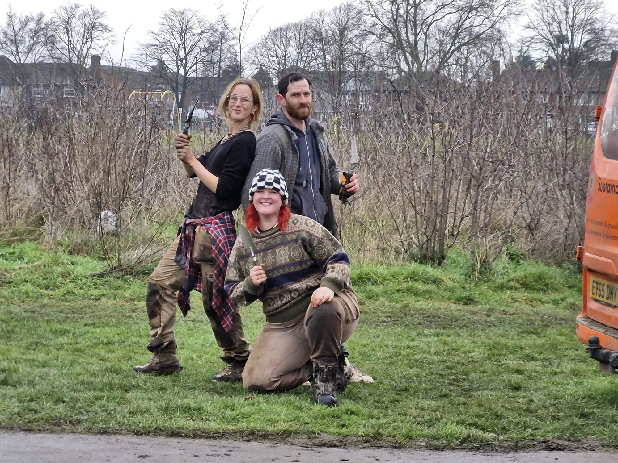 Two people pose playfully outdoors, with one crouching beside a dog in a grassy area. A vehicle is partially visible.