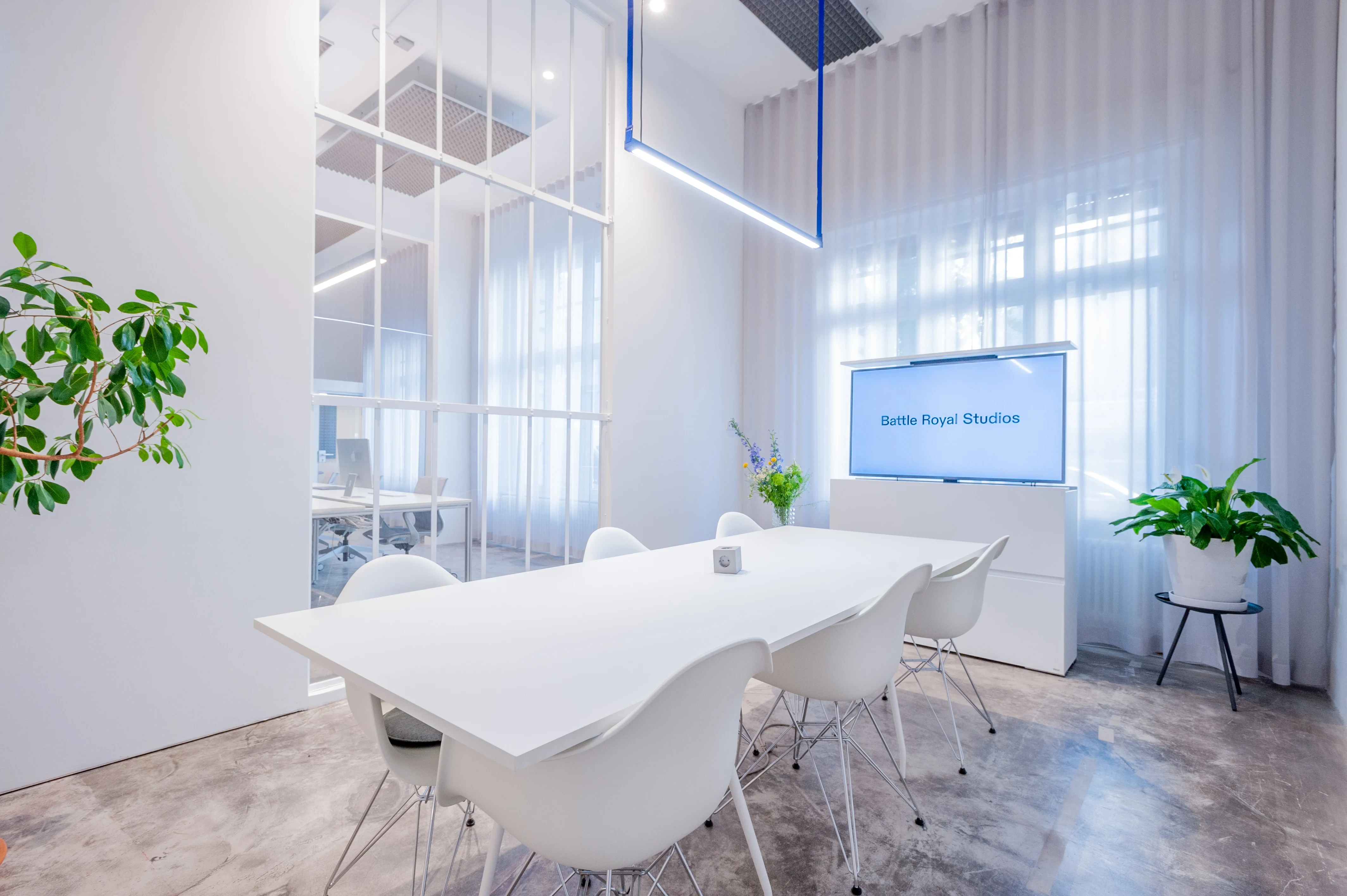 White meeting room at BATTLE ROYAL with a long white table, white Eiffel-base chairs, a screen reading Battle Royal Studios on a white sideboard, plants and a cobalt-blue pendant frame above.