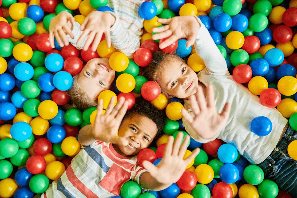 Three children laugh while lying in a colorful ball pit filled with red, blue, yellow, and green plastic balls, playfully reaching towards the camera.