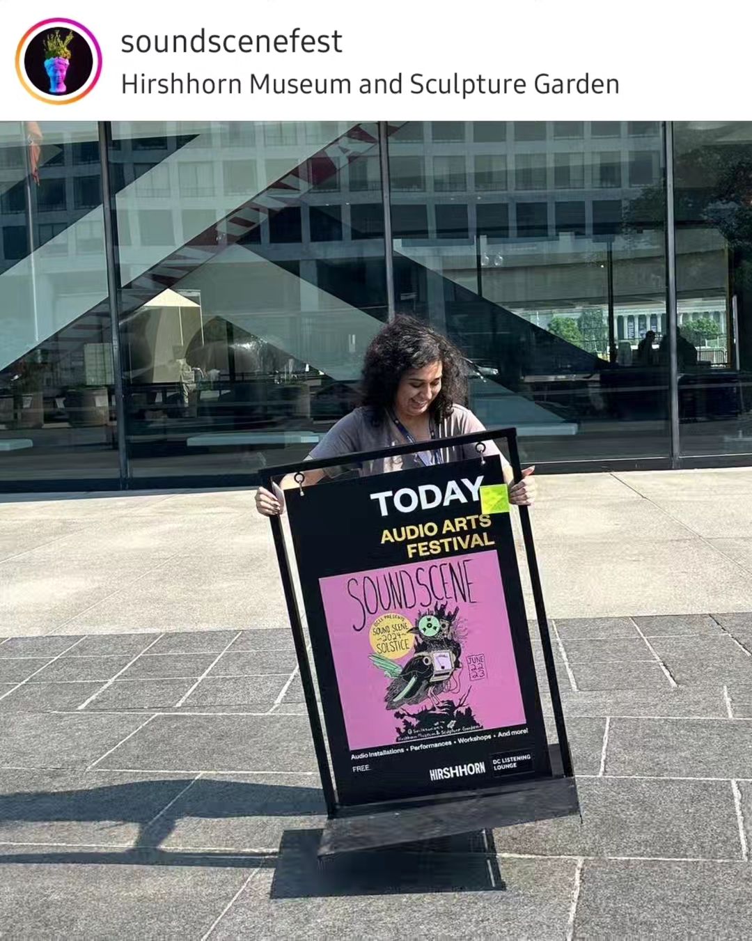 A person with curly hair and a lanyard smiles while holding a large event sign outside the HIrshhorn Museum building. The sign promotes the 'Sound Scene' Audio Arts Festival at the, featuring a pink poster with an illustrated design.