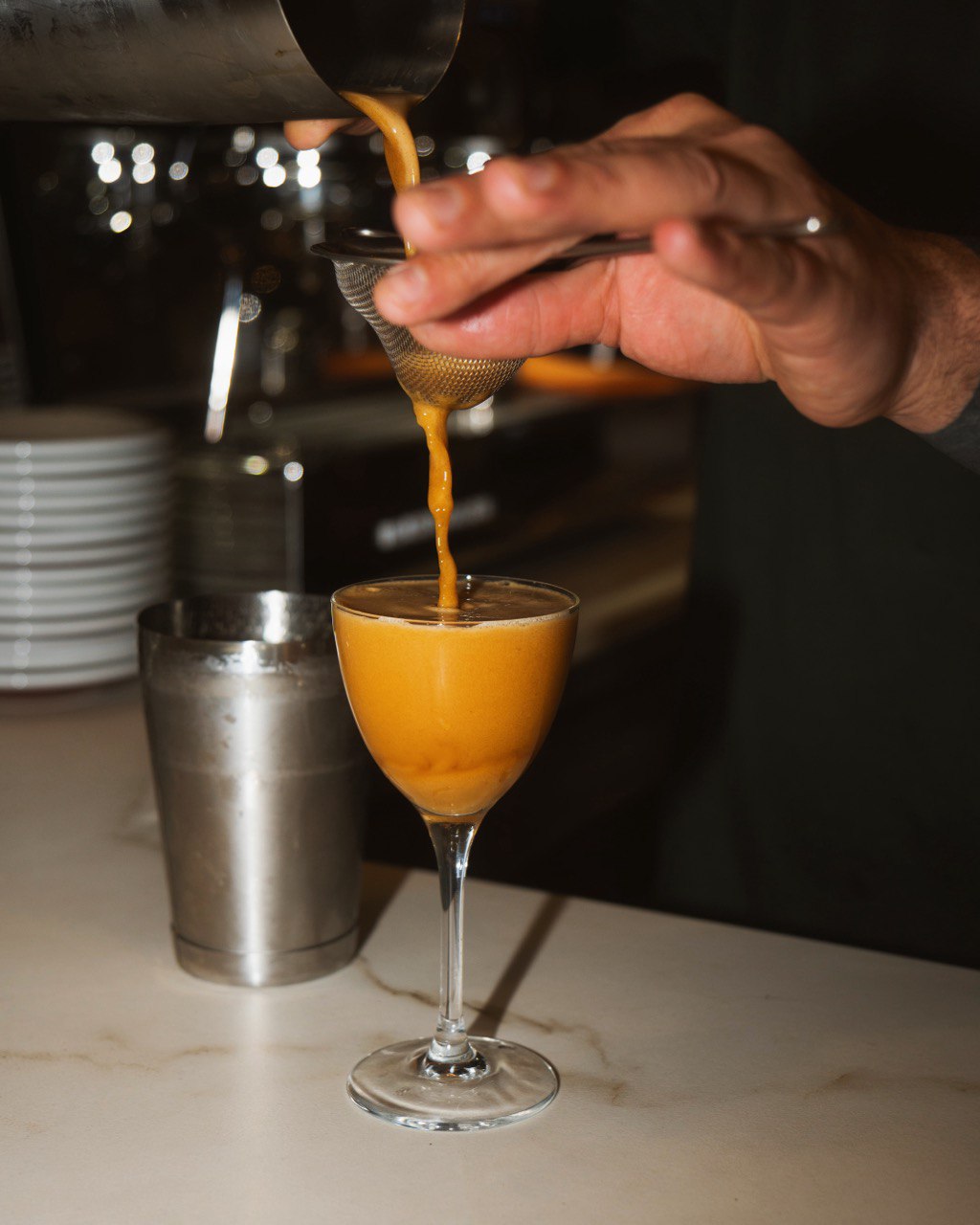 A bartender is pouring a freshly shaken orange cocktail through a fine strainer into a stemmed glass on a marble countertop, with a cocktail shaker and stacks of white plates in the blurred background.