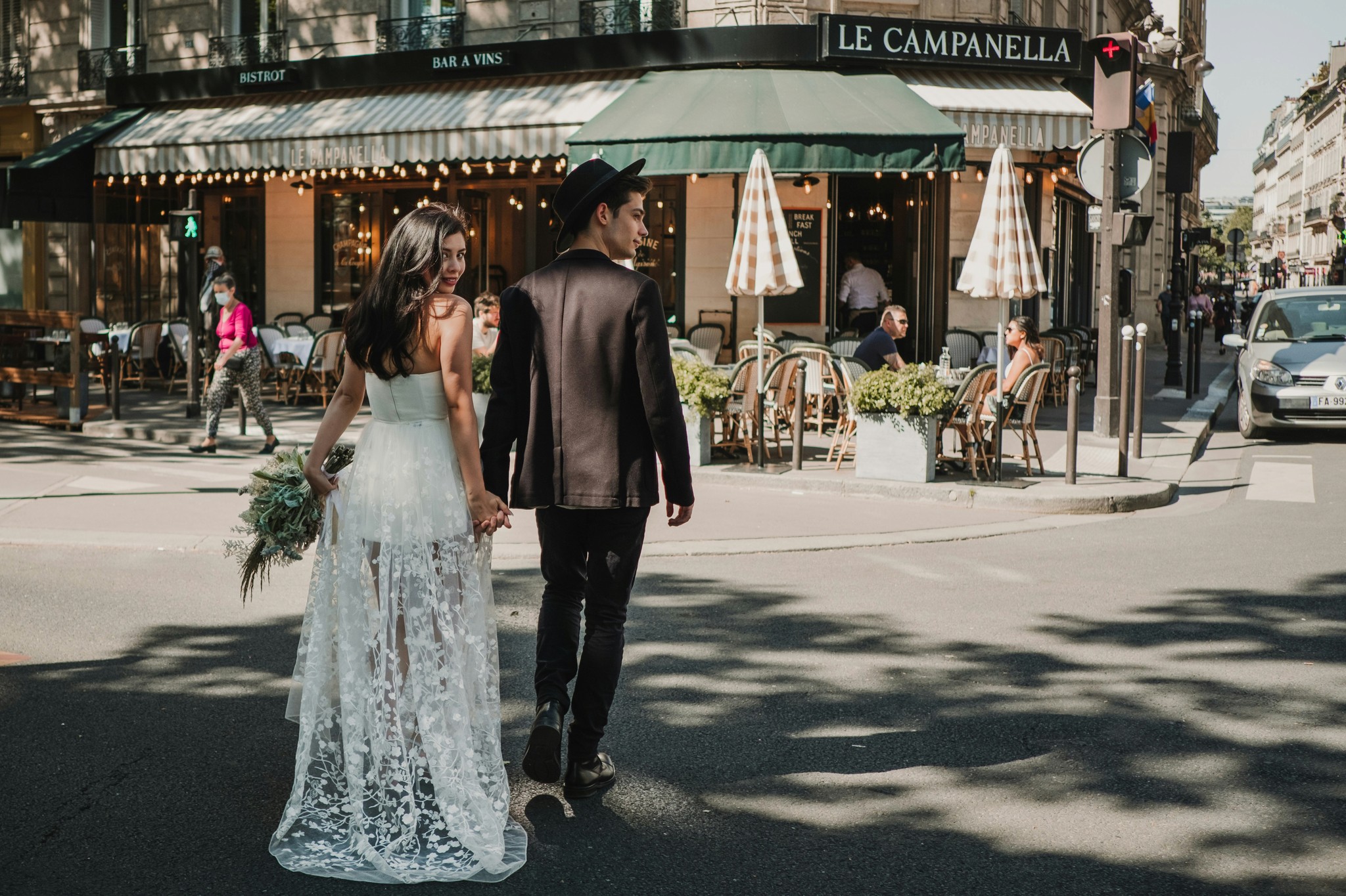 Couple strolling through a sunlit Paris street corner with the bride holding a bouquet and her lace dress trailing behind